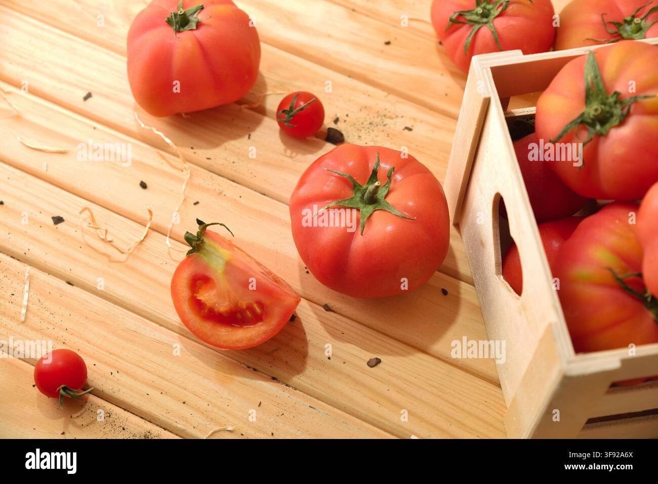 Pomodori maturi riposano su una superficie di legno illuminata dal sole accanto a una cassa rustica, con un pomodoro affettato che rivela il suo interno succoso e i semi. Foto Stock