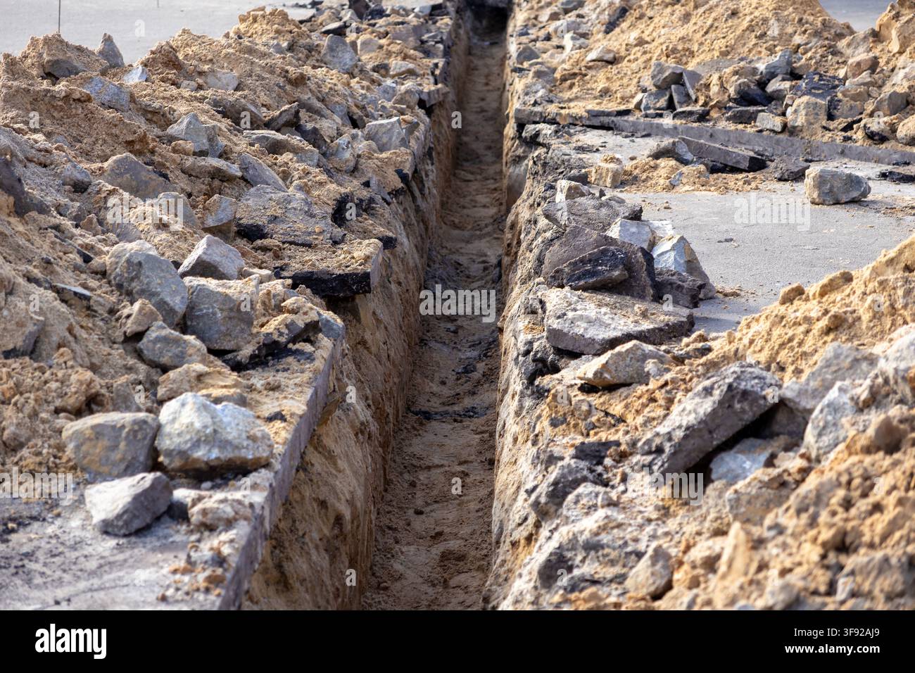 Un canale aperto scavato per la manutenzione delle utenze sotterranee, con pile di detriti e terra sciolta lungo una strada suburbana per simboleggiare i lavori in corso Foto Stock