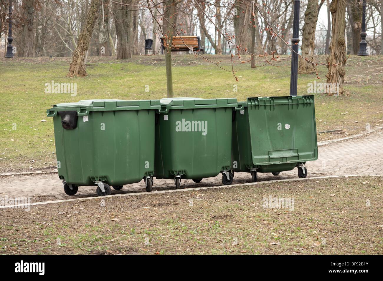 Grandi contenitori di spazzatura in plastica sono allineati lungo un sentiero in un giardino boscoso, rappresentando una manutenzione ambientale municipale efficiente e organo Foto Stock