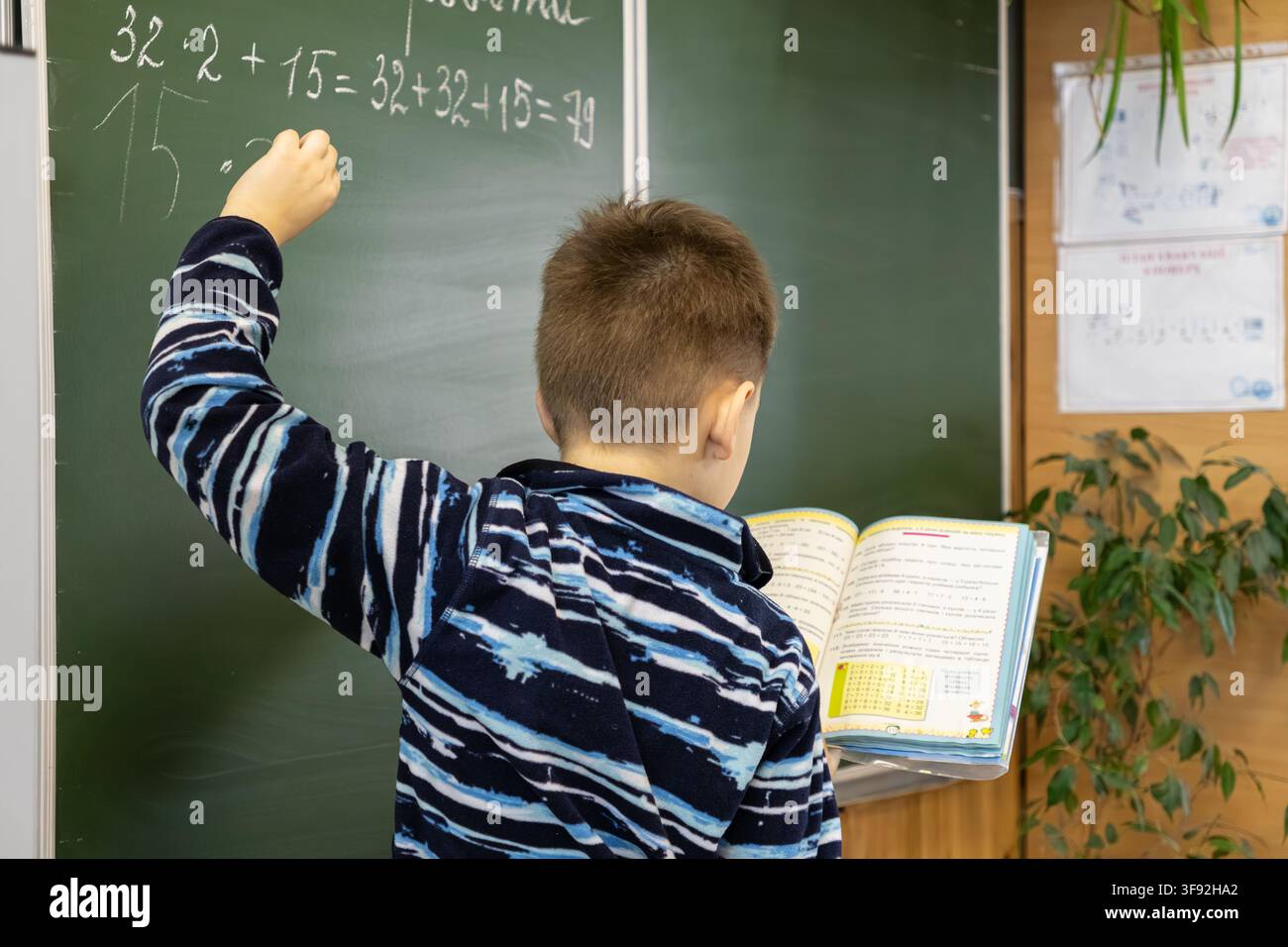 Kyiv, Ucraina - 20 marzo 2026: Uno studente concentrato risolve difficili problemi di matematica su una lavagna in classe utilizzando un libro di riferimento per illustrare il suo Foto Stock