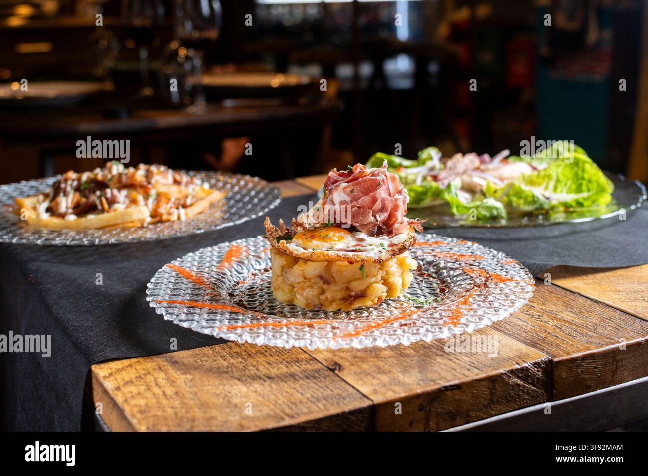 Piatto di patate in stile spagnolo sormontato da uovo fritto e prosciutto stagionato, servito su un piatto elegante con altri piatti in sottofondo. Foto Stock