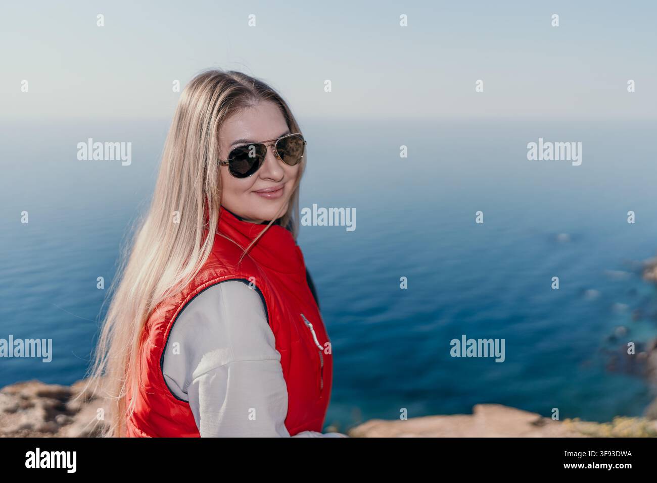 Donna viaggio mare Bionda in giubbotto rosso e occhiali da sole che gode di una vista panoramica sulla costa in una giornata soleggiata con spazio per il testo Foto Stock