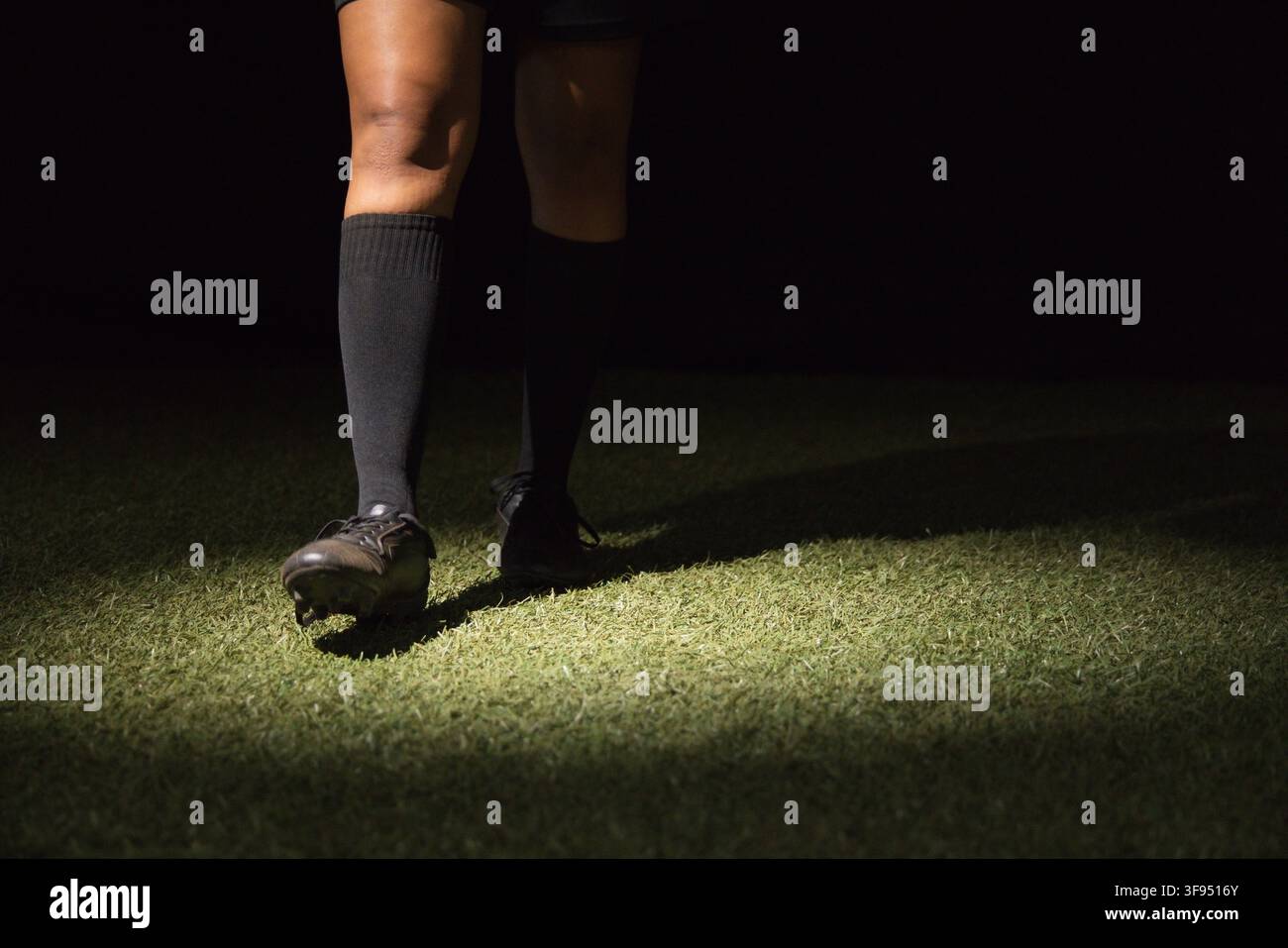 Giocatrice di calcio in piedi su erba sintetica di notte, indossa calzini al ginocchio e scarpette da calcio sotto i riflettori Foto Stock