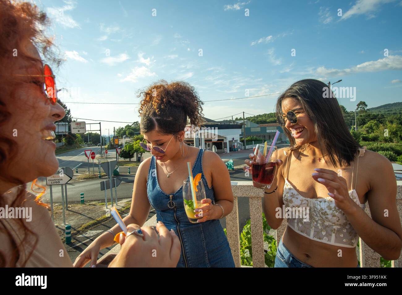 Tre giovani donne che ridono con bevande colorate su un balcone soleggiato accanto a una strada rurale, Foto Stock