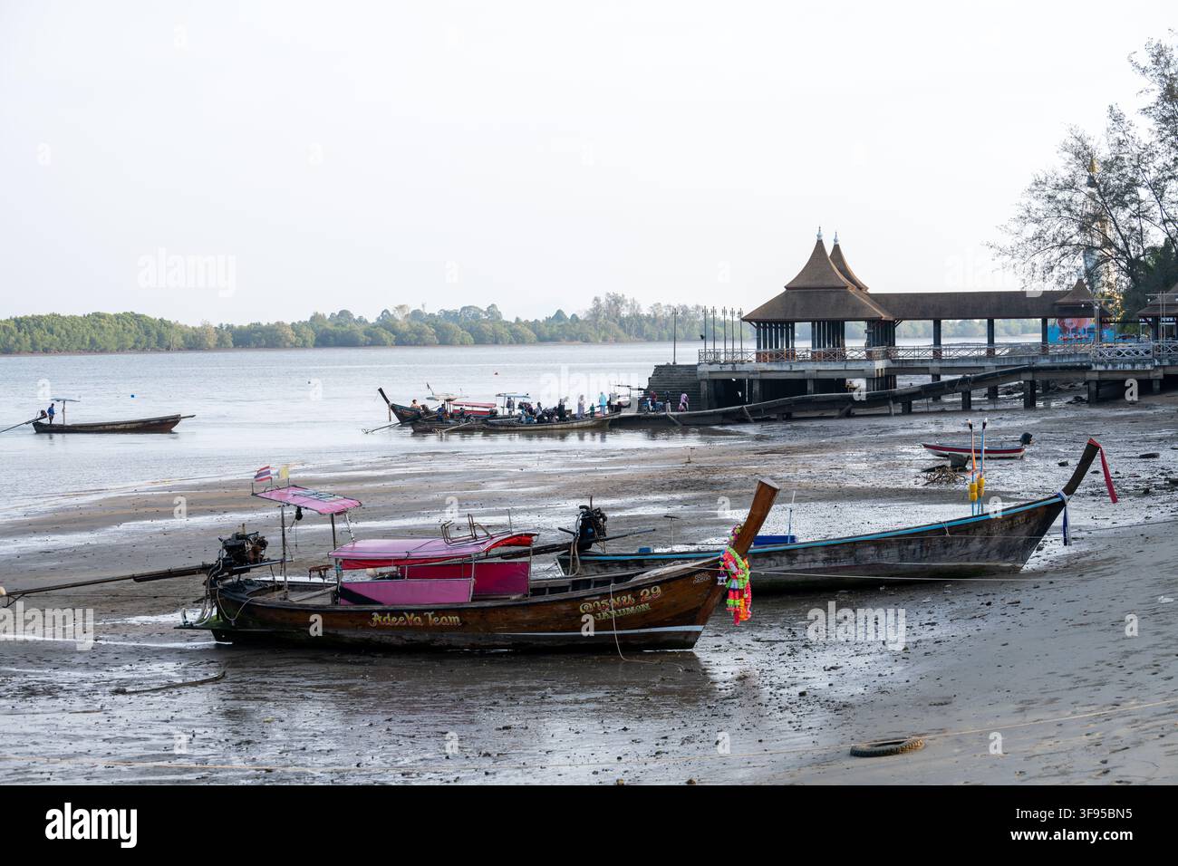 KRABI, THAILANDIA — 4 MARZO 2026: Barche a lunga coda si riposano sulla riva esposta alle maree mentre passeggeri e operatori di barche si radunano accanto a un molo sul lungomare. Foto Stock