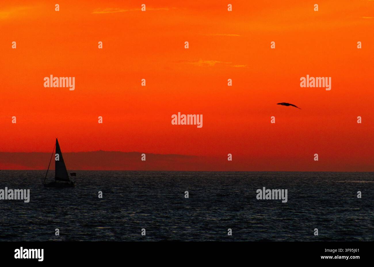 Barca a vela scivola su un oceano calmo al tramonto infuocato. Un caldo bagliore arancione circonda il sole che si immerge nell'orizzonte. La scena evoca pace, solitudine e un'arrivo tranquilla Foto Stock