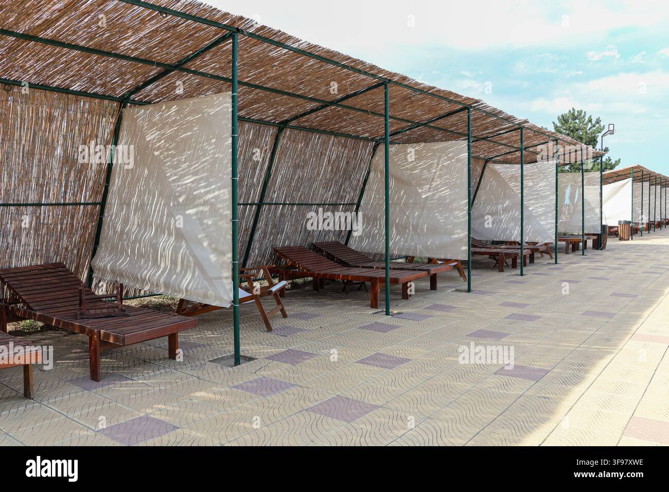 Sdraiini in legno disposti sotto una copertura di canne in un resort estivo, con luce solare a motivi, ombra e una forte prospettiva attraverso l'area di relax all'aperto Foto Stock