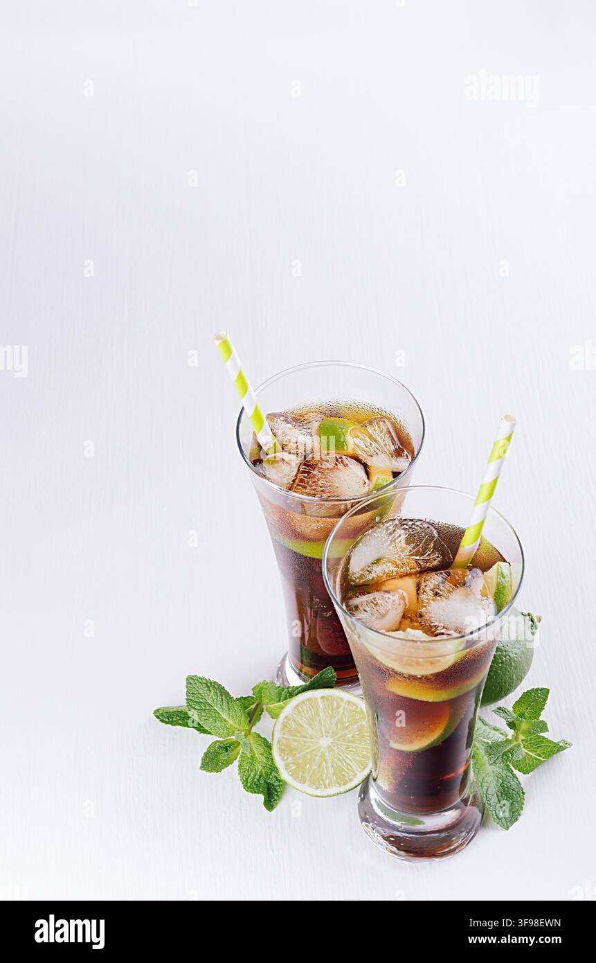 Cocktail freddo cuba libre in due bicchieri bagnati con paglia, pezzi di lime, menta su morbido sfondo bianco moderno, vista dall'alto, bordo. Foto Stock