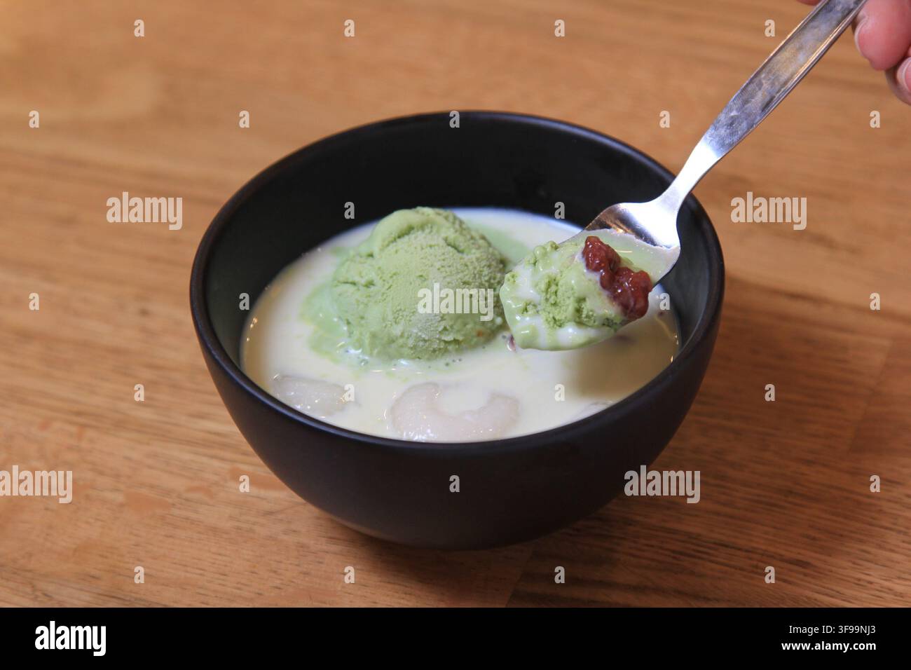 Prima vista di un cucchiaio di gelato al tè verde matcha con fagioli rossi in una piccola ciotola nera, tenuto da un cucchiaio, su un dessert cremoso con litchi Foto Stock