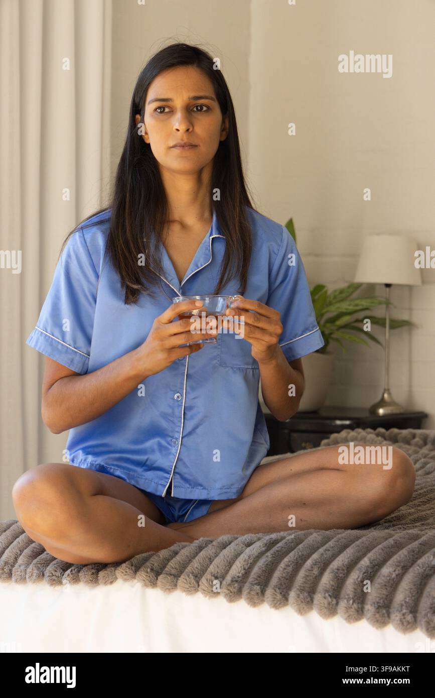 Donna adulta seduta a gambe incrociate sul letto, indossa un pigiama azzurro e tiene in mano una tazza da tè di vetro trasparente Foto Stock