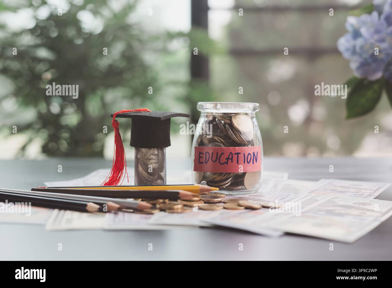 Barattolo di risparmio per l'istruzione pieno di monete accanto a una banconota arrotolata sormontata da un berretto accademico, con matite e documenti finanziari che simboleggiano le tasse universitarie Foto Stock