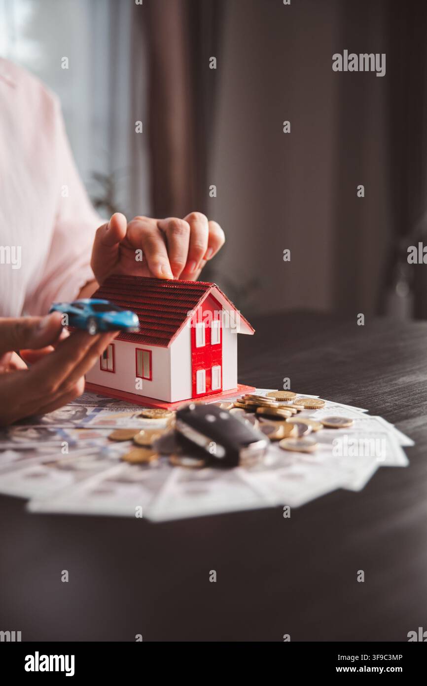 Una persona indica una macchina bianca, pile di monete in aumento e una casa con tetto rosso, rappresentando la strategia di risparmio, la pianificazione del prestito, l'acquisto di proprietà e il finanziamento del veicolo Foto Stock