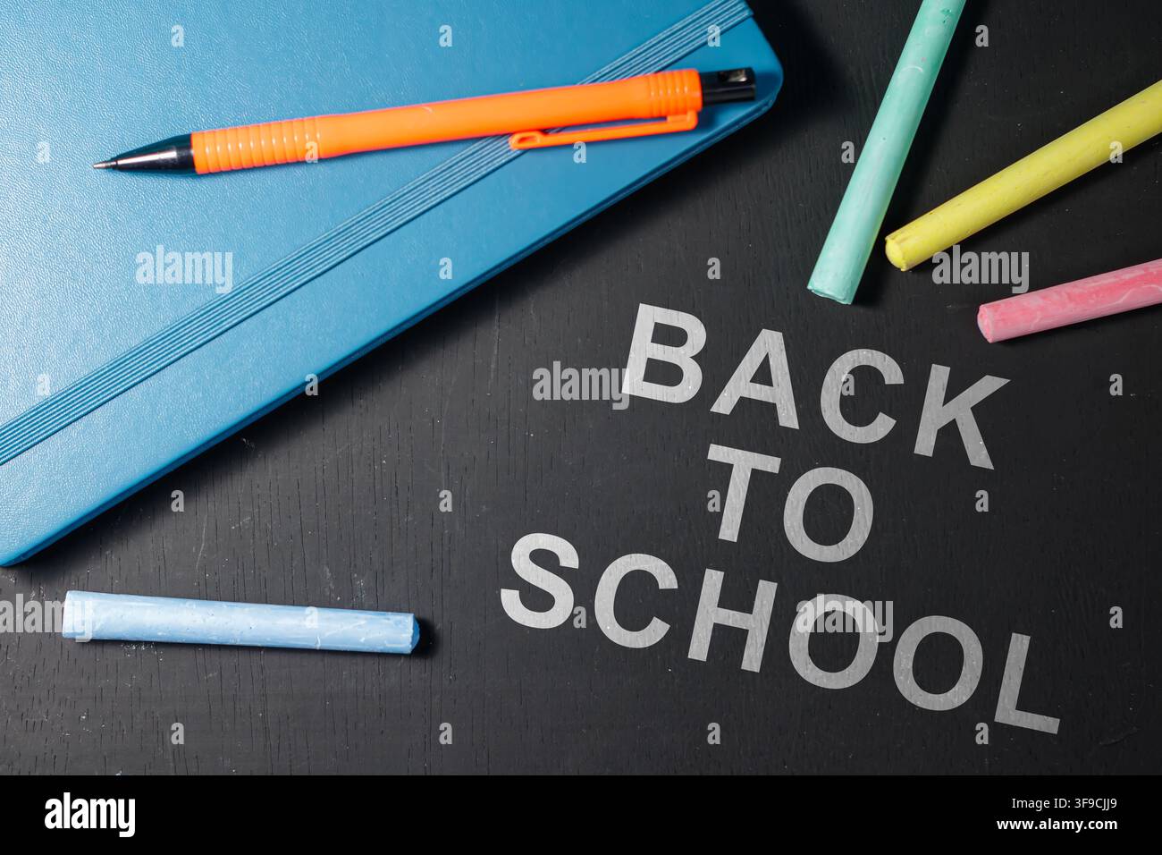 Messaggio di ritorno a scuola su una superficie nera circondata da gessi colorati e un quaderno blu con una matita arancione sopra. Foto di alta qualità Foto Stock