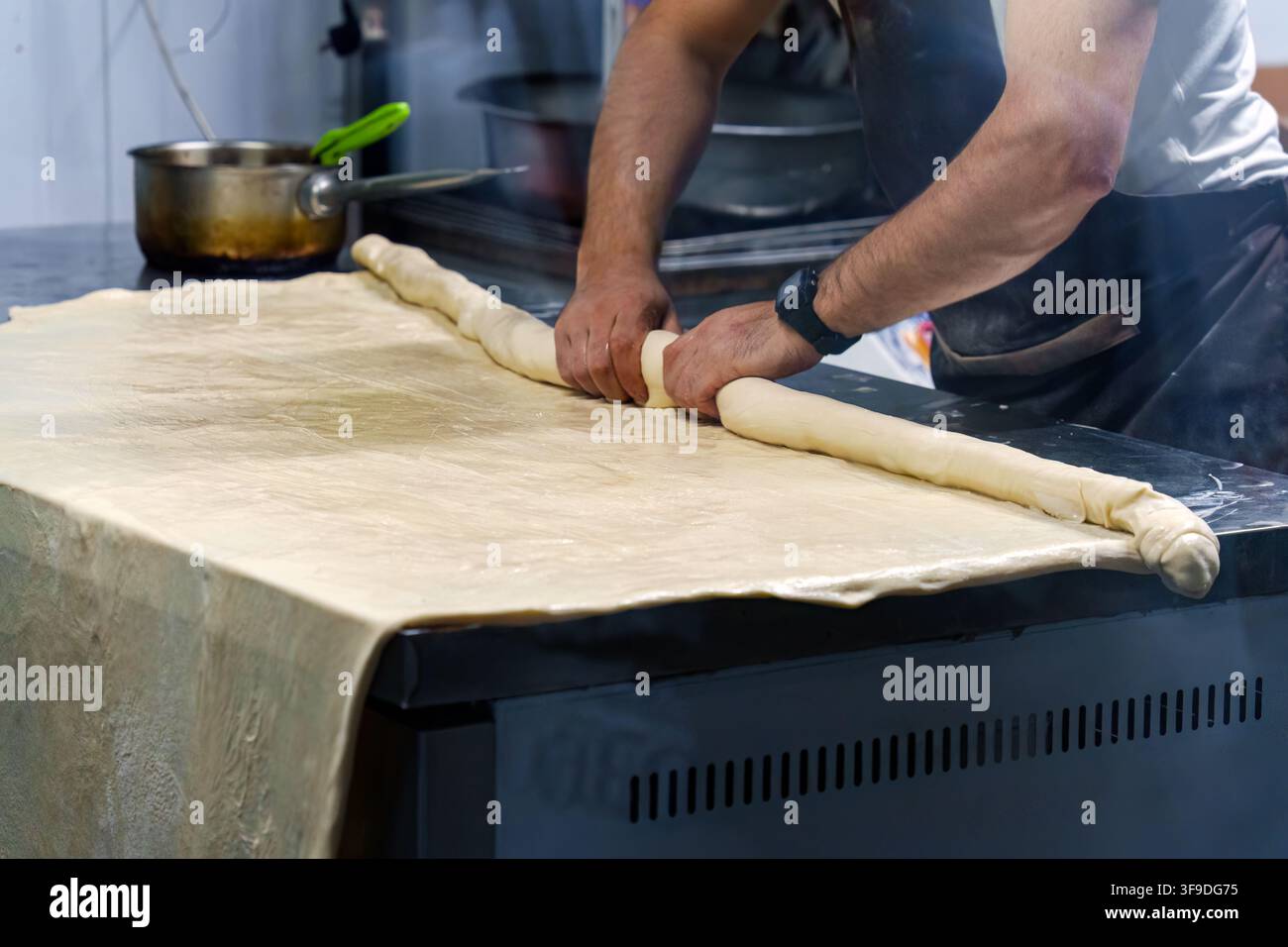 Panettiere esperto che arrotola una sfoglia di pasta in forma di tronco su un grande bancone in cucina. Artigianato tradizionale di produzione del pane con le mani che modellano la pasta fresca in Foto Stock
