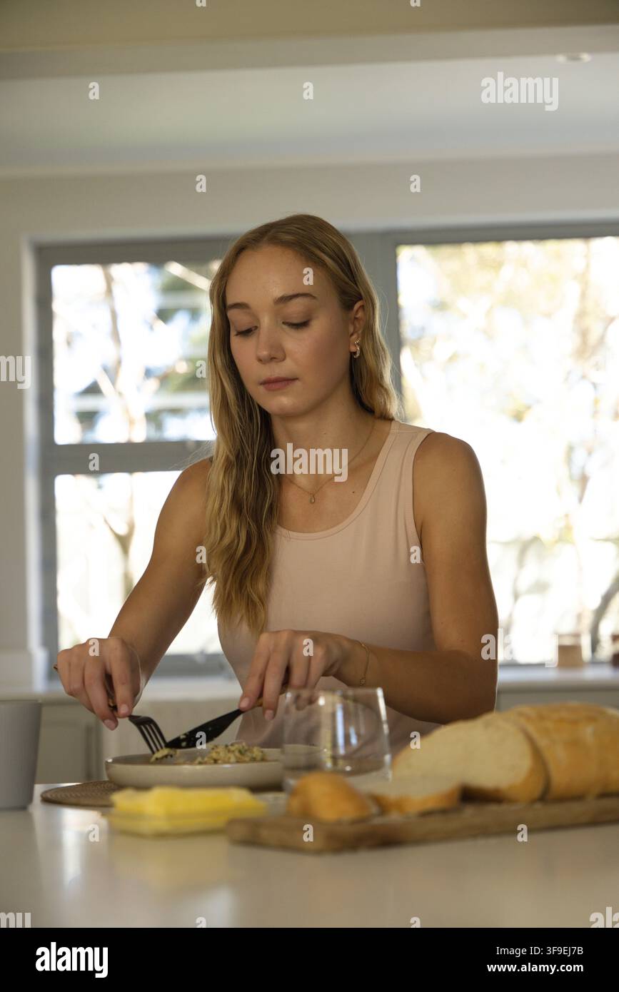 Donna sulla ventina con canottiera seduta al bancone vicino alla finestra che usa forchetta e coltello sopra una ciotola Foto Stock