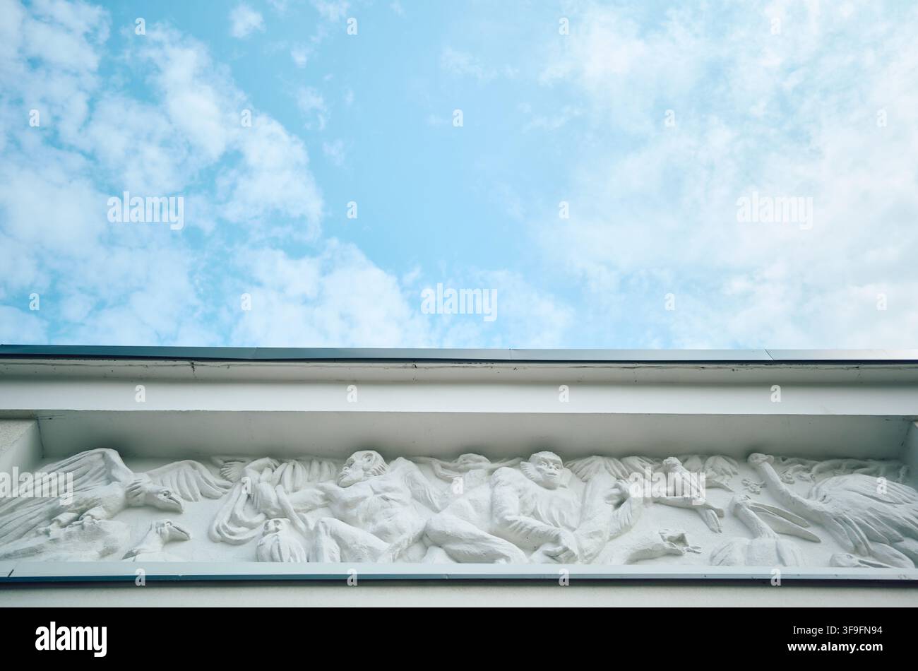 Rilievo architettonico a fregio con figure scolpite e creature alate sotto la cornice, visto da un angolo basso contro un ampio cielo blu pallido con morbido Foto Stock