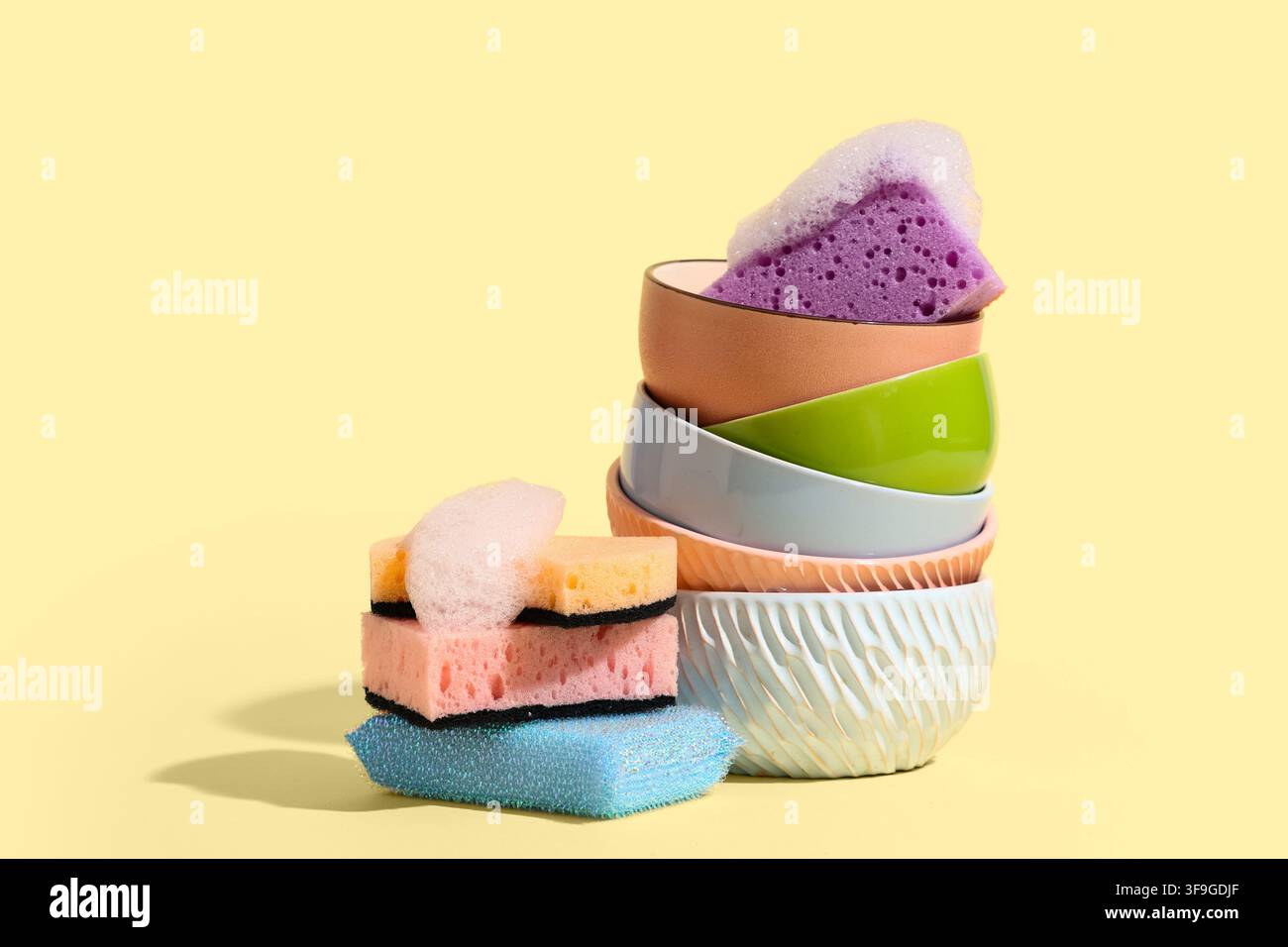 Pila di ciotole in ceramica colorate con spugne per lavare i piatti e schiume di sapone su uno sfondo giallo, Pulizia della cucina Foto Stock