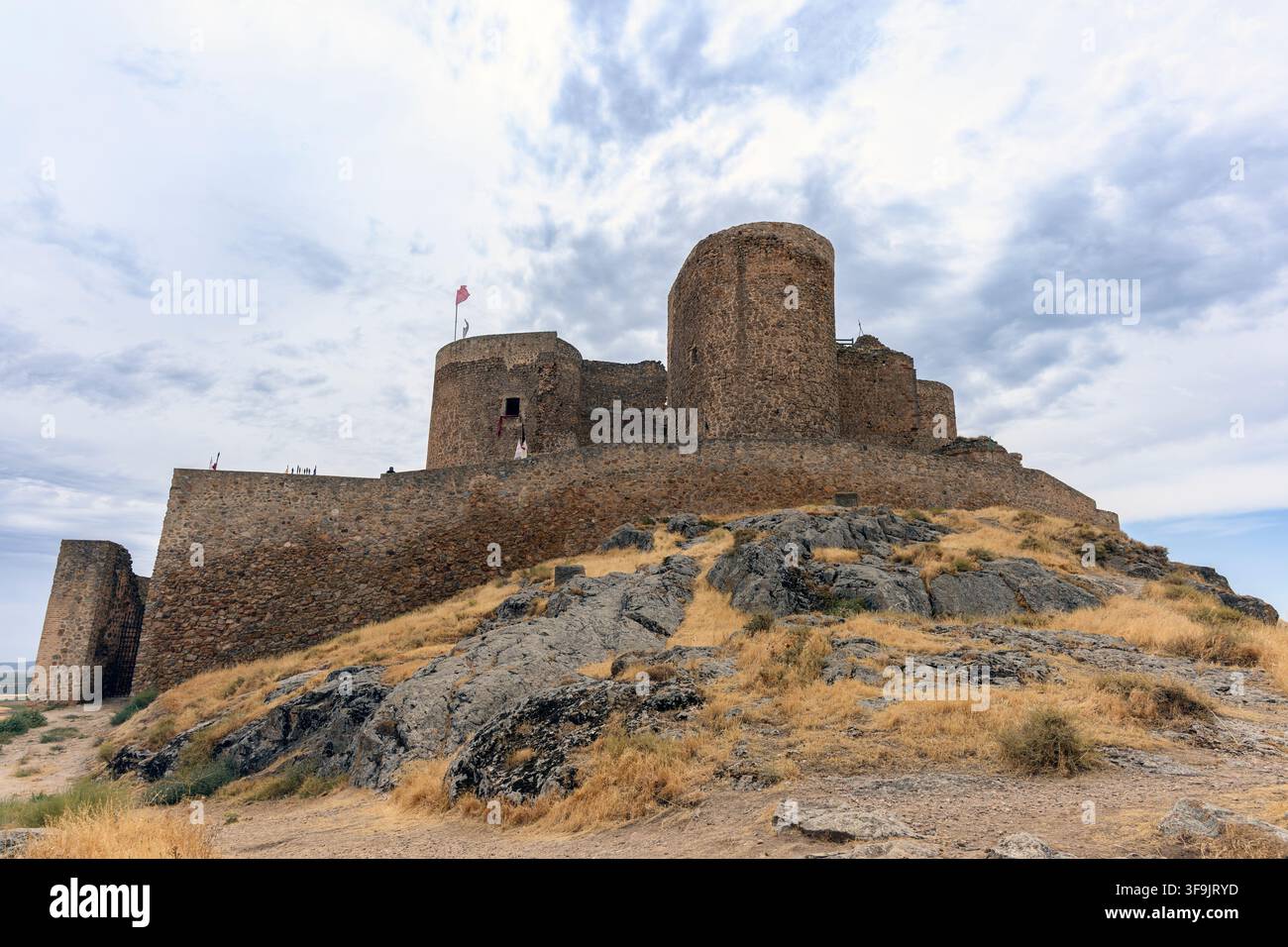 Muri di pietra coprono il pendio. Torri si ergono dal terreno roccioso. Una bandiera sventola sulla cima del castello. Erba resistente alla siccità punteggia il pendio arido. Nuvola Foto Stock