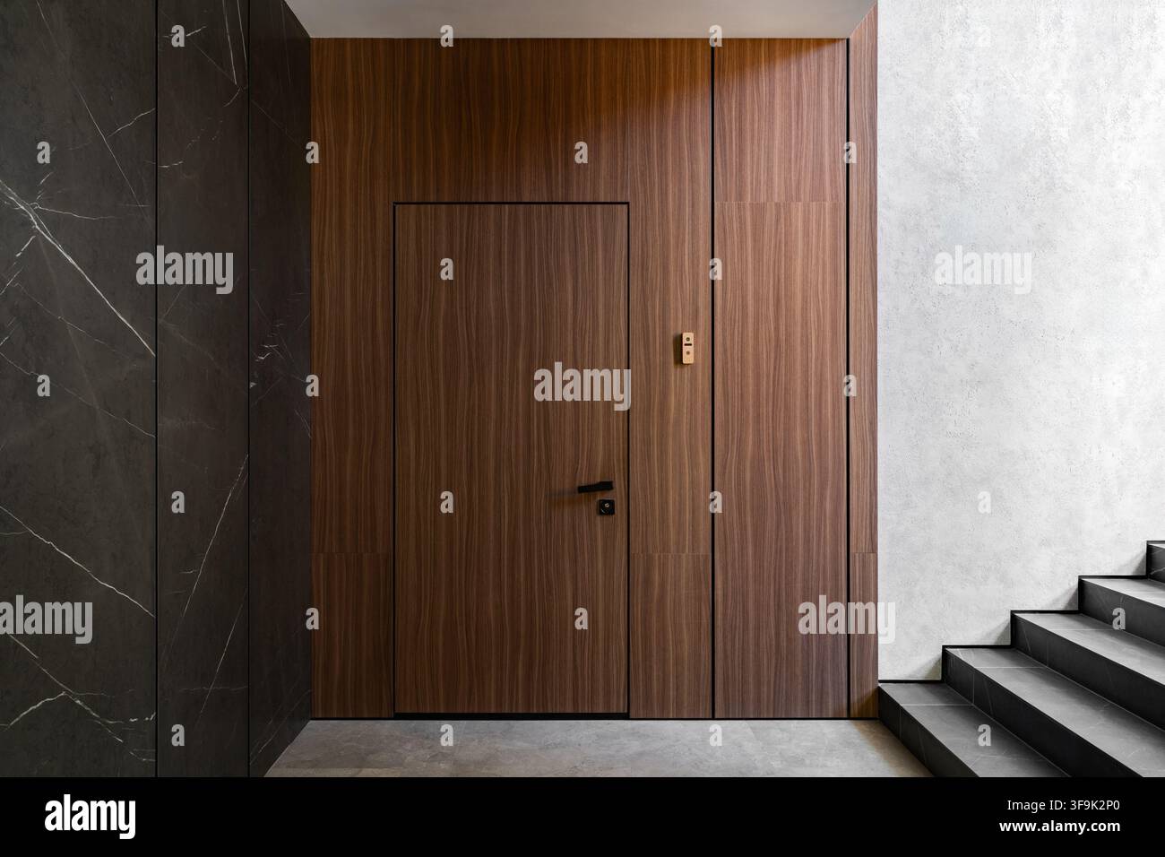 Porta d'ingresso di un appartamento di lusso in un complesso residenziale esclusivo con porta principale rivestita in noce, pareti effetto marmo scuro e scala minimalista Foto Stock
