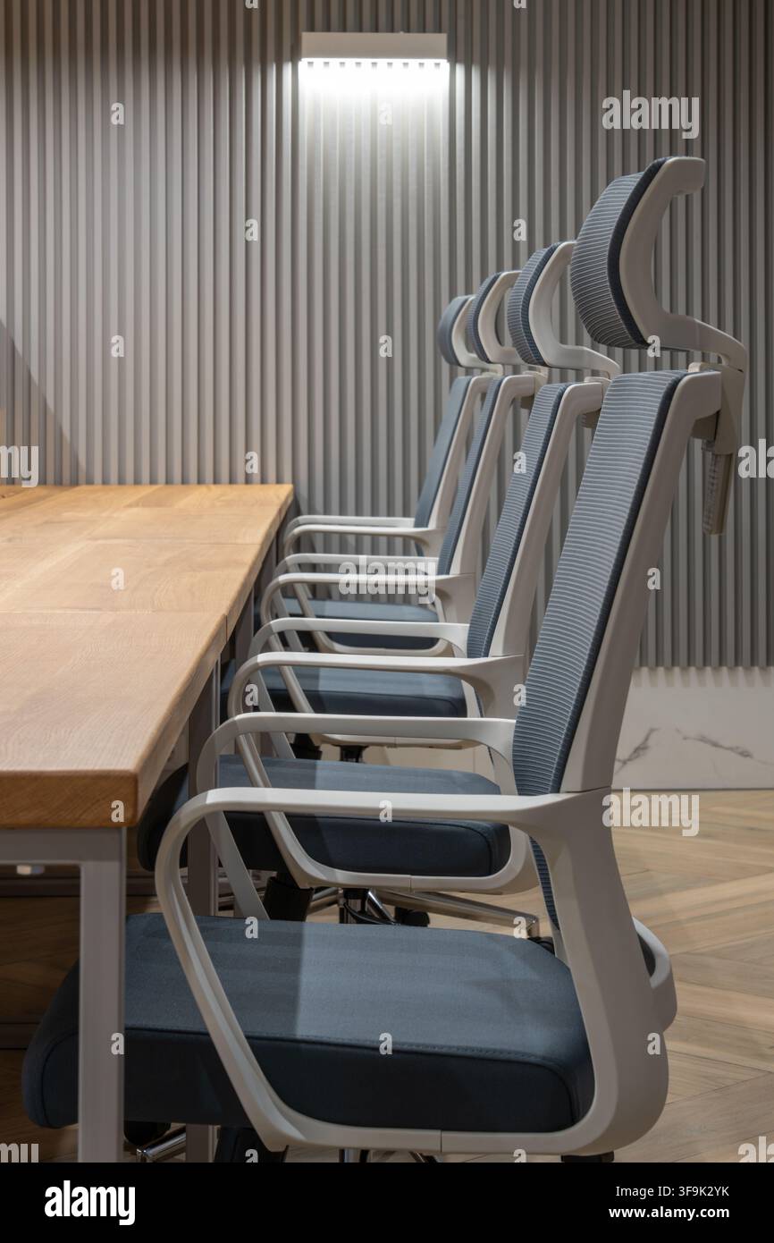 Interno di ufficio contemporaneo con sedie da conferenza ergonomiche disposte lungo un tavolo riunioni in legno, parete a doghe acustiche e lampada da parete minimalista Foto Stock
