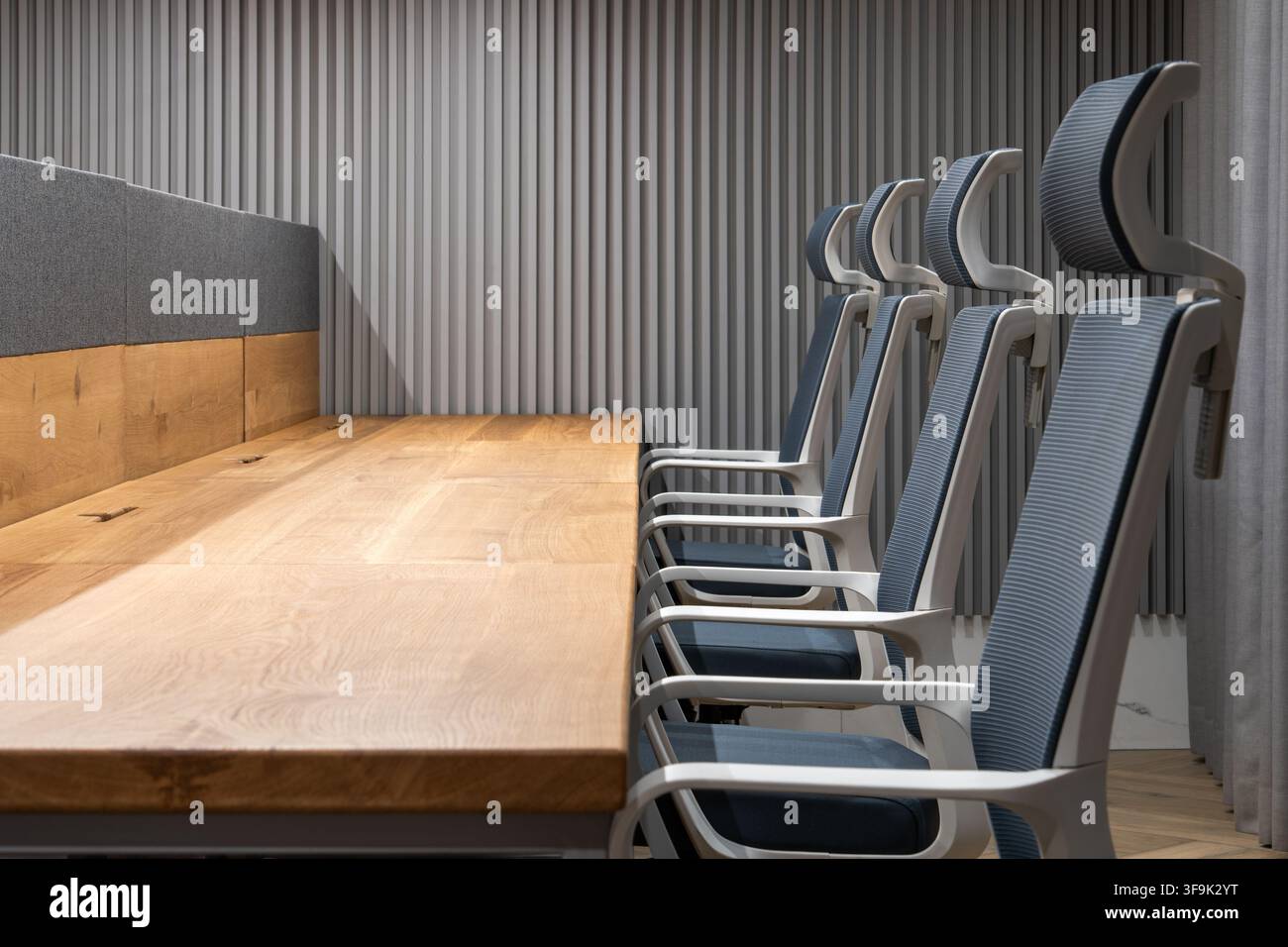 Interno di ufficio contemporaneo con sedie da conferenza ergonomiche disposte lungo un tavolo riunioni in legno, parete a doghe acustiche e lampada da parete minimalista Foto Stock