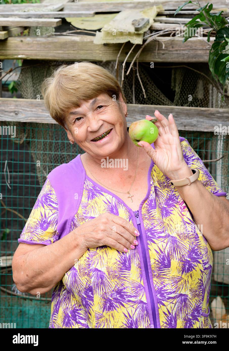 Allegra donna anziana russa di 73 anni sorride e tiene una mela fresca in un cortile rurale. Foto Stock