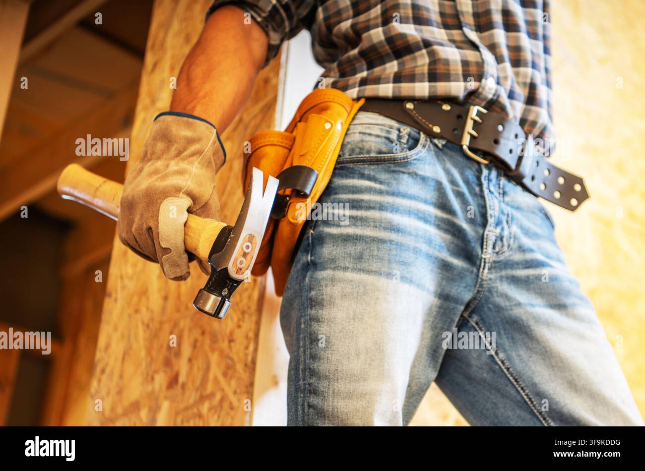 Una persona si trova in un cantiere edile tenendo un martello in una mano. L'individuo indossa guanti e ha una cintura degli attrezzi. Pareti e materiali di legno sono v Foto Stock
