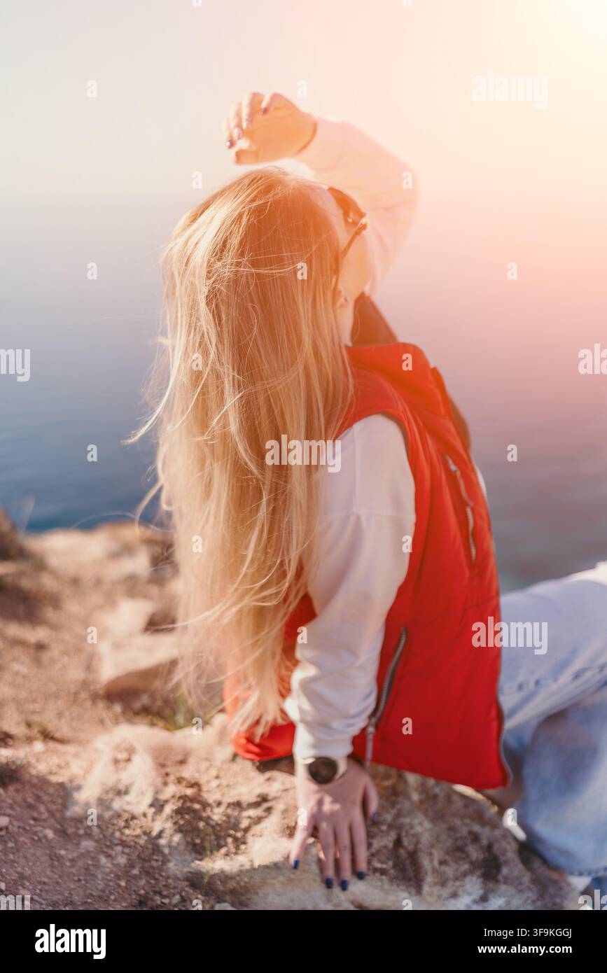 Donna bionda in giacca rossa seduta su una scogliera rocciosa in riva al mare durante un tramonto luminoso e soleggiato con flare dell'obiettivo Foto Stock
