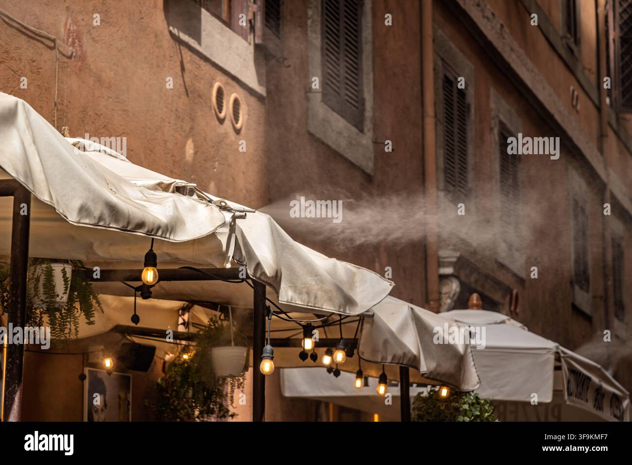 Un ristorante utilizza ugelli di spruzzatura a soffitto per spruzzare una fine nebbia rinfrescante sui commensali all'aperto durante un'ondata di caldo a Roma, Italia. Foto Stock