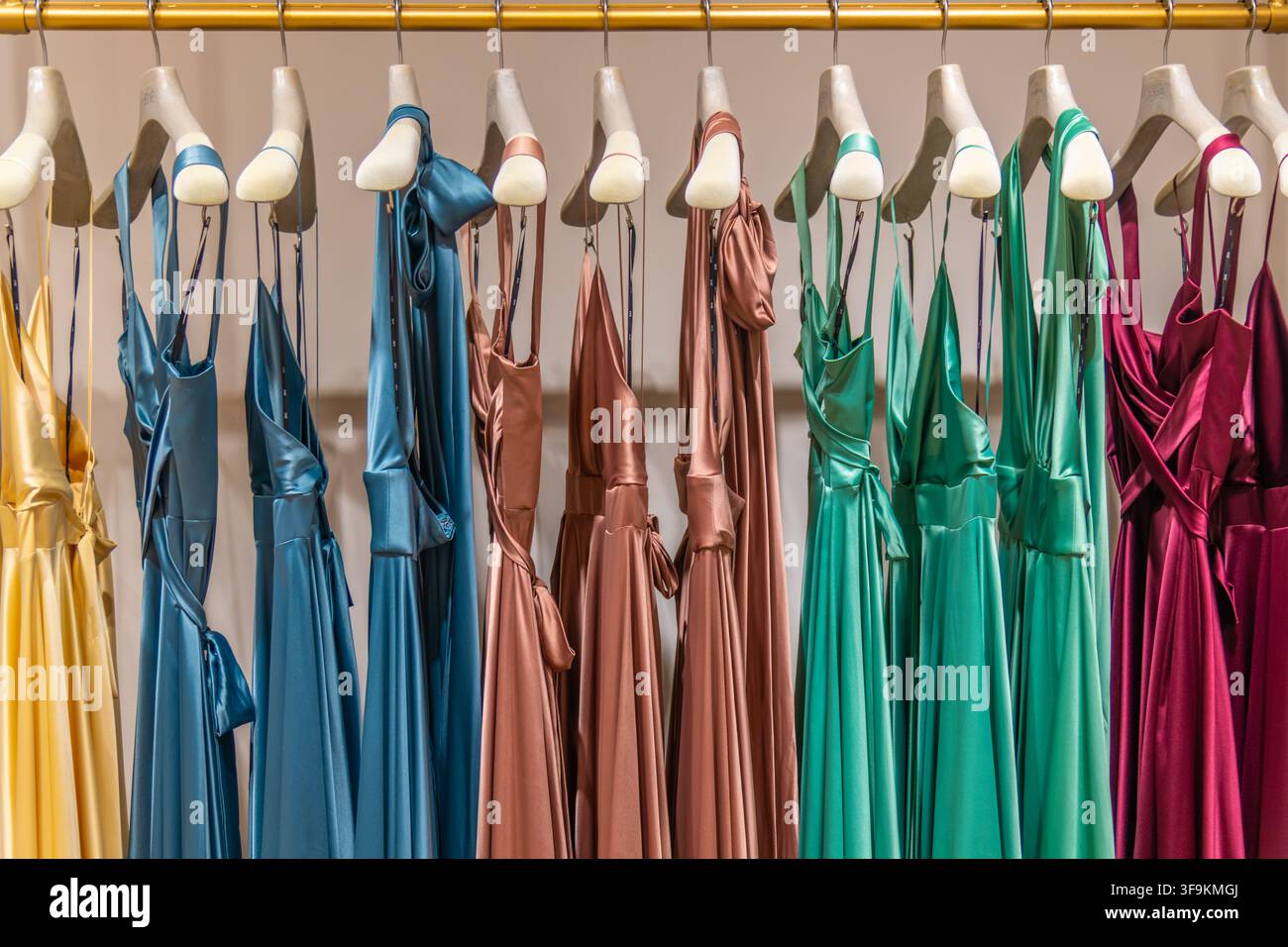 Una fila di abiti di raso colorati in senape, blu, rame, verde e bordeaux è appesa a grucce di legno lungo una rastrelliera di abiti di una boutique. Foto Stock