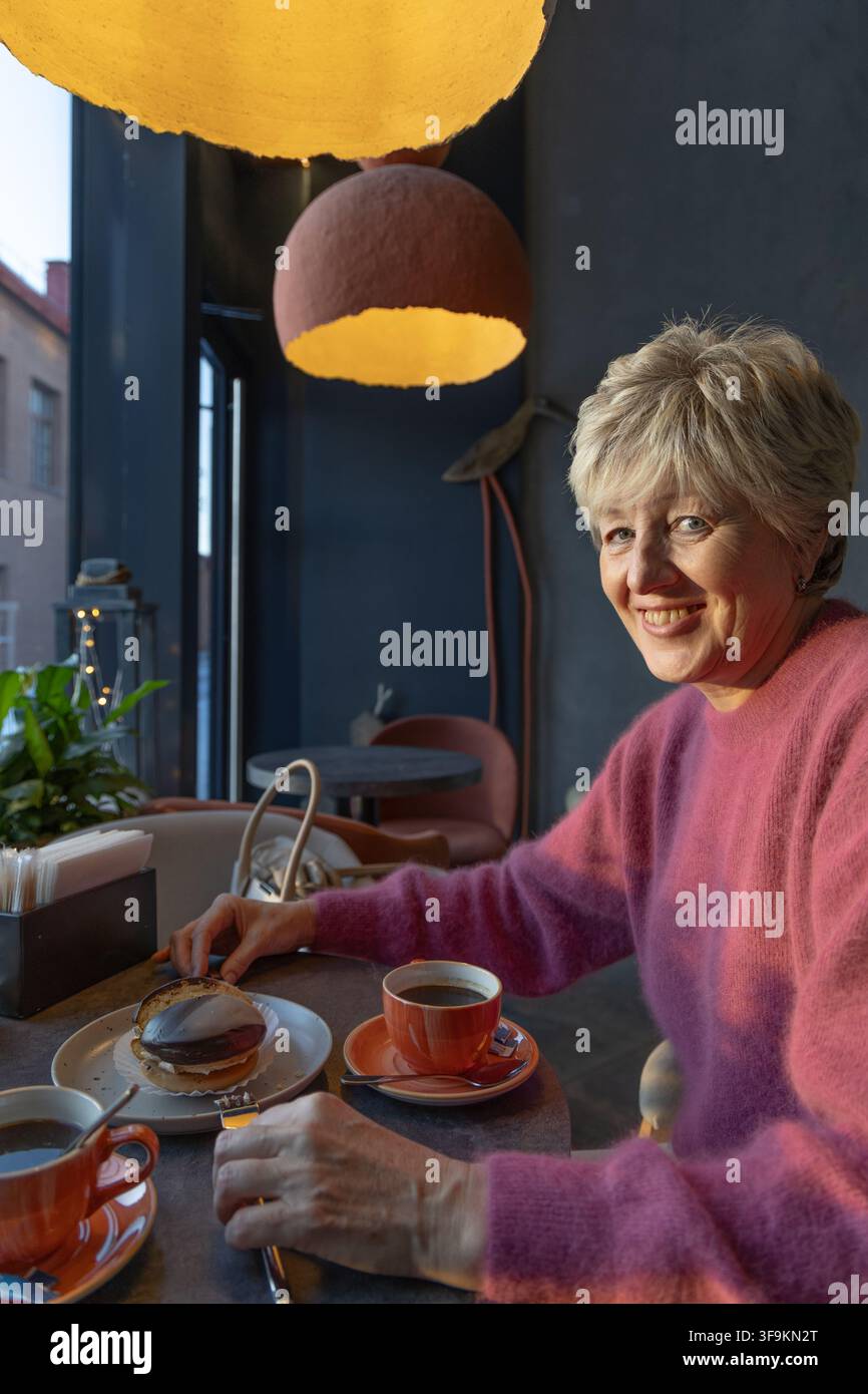 Elegante donna matura con una bellezza senza tempo che sorride alla telecamera mentre gusta caffè e pasticcini in un moderno caffè scandinavo. Foto Stock