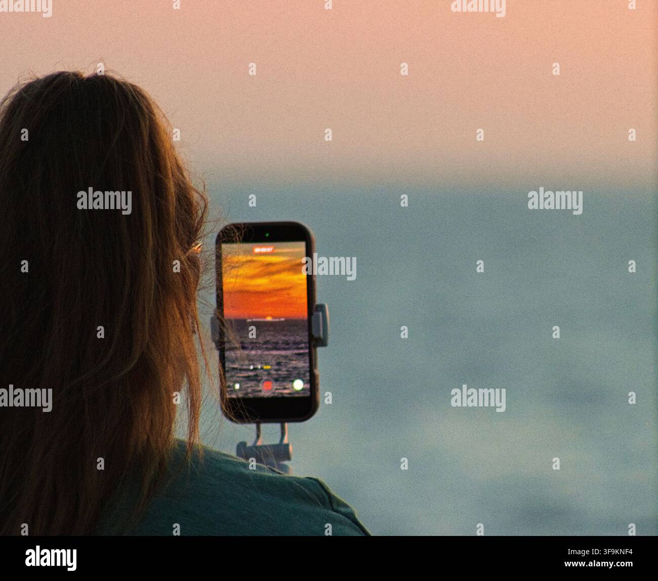 Una persona cattura il tramonto sull'oceano con il telefono. La vista da dietro mostra un momento concentrato e calmo. Ideale per temi di viaggio, tecnologia o natura. Perfetto per Foto Stock