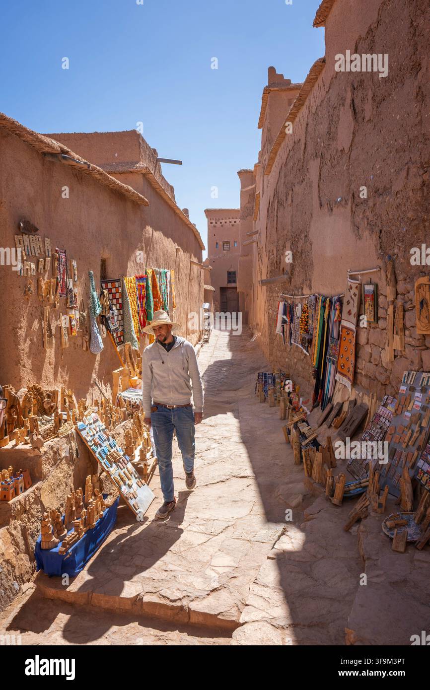 Uomo che cammina lungo una stretta via mercatale fiancheggiata da souvenir di artigianato locale e tappeti contro muri in mattoni di argilla rossa sotto la luce intensa del sole ad Ait B Foto Stock