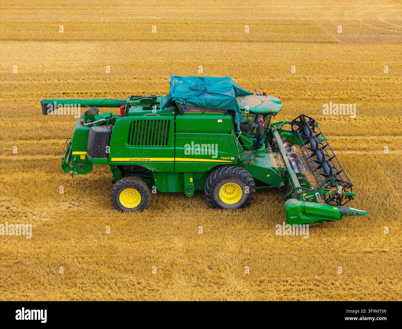 Kazupe, Lettonia - 01 agosto 2026: Vista aerea di una mietitrebbia rotta parcheggiata in un vasto campo di grano dorato, che mostra dettagli meccanici e un blu Foto Stock