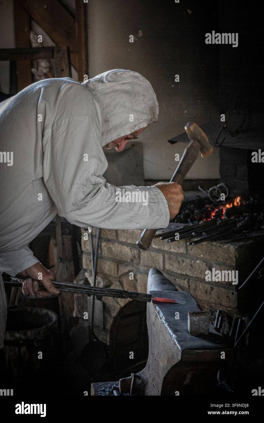 Rievocazione di un fabbro medievale nel centro di Sandwich – in costume, aziona il mantice con metallo rovente in pinze vicino al fuoco nella sua bottega Foto Stock