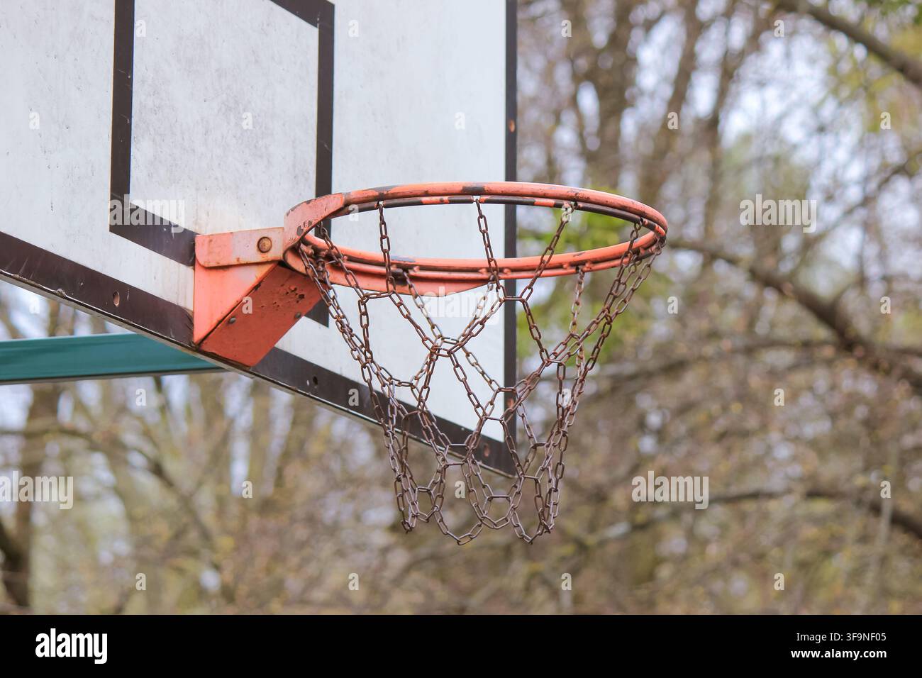 Primo piano di un canestro da basket logoro con una rete a catena resistente in un contesto di parco pubblico Foto Stock