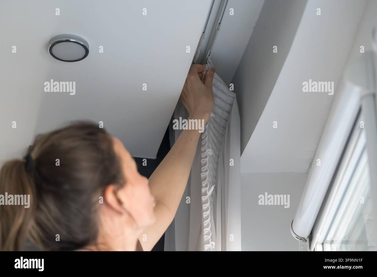 Primo piano di una donna su una scala a pioli che appende una tenda bianca di garza (tulle) appena lavata su un'asta per tende accanto a una finestra. Pulizia di primavera, faccenda domestica. Foto Stock