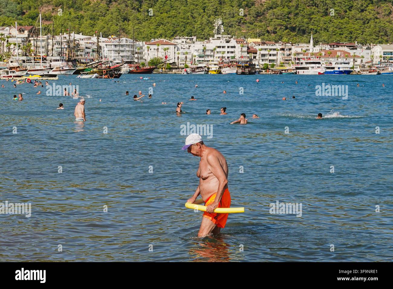 Uomo anziano in pantaloncini arancioni che tiene una galleggiante a forma di spaghetti cammina nell'acqua del mare con persone che nuotano e la città di Marmaris sullo sfondo, Turchia. Foto Stock