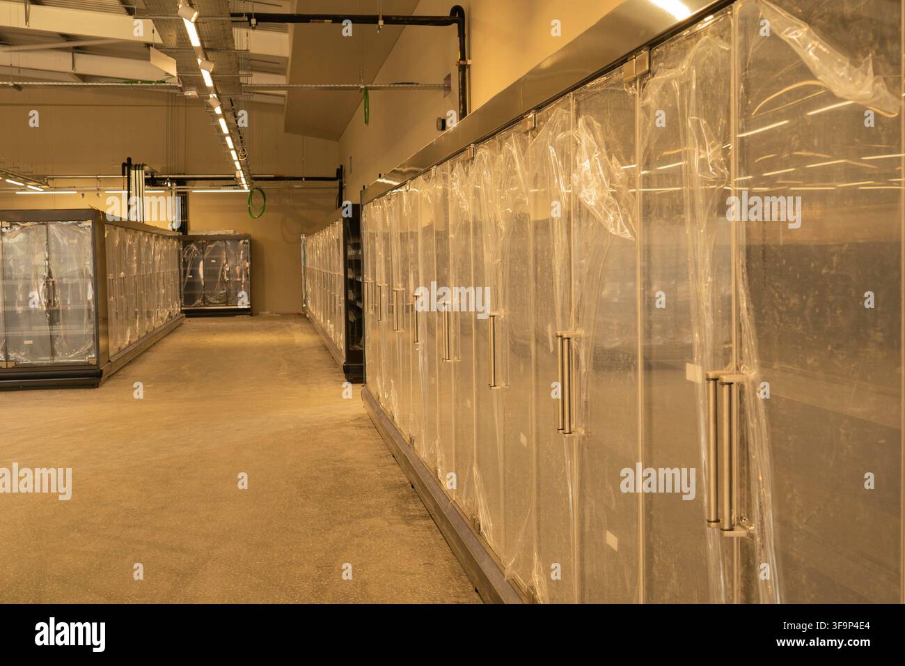 File di armadi frigoriferi commerciali con porte in vetro nuovi di zecca coperti da pellicola protettiva all'interno di un supermercato non finito in costruzione Foto Stock