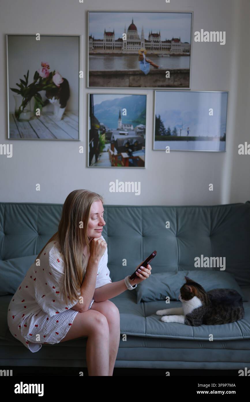 Giovane donna che usa lo smartphone seduta sul divano a casa con il suo gatto, concetto di stile di vita digitale quotidiano. Foto Stock