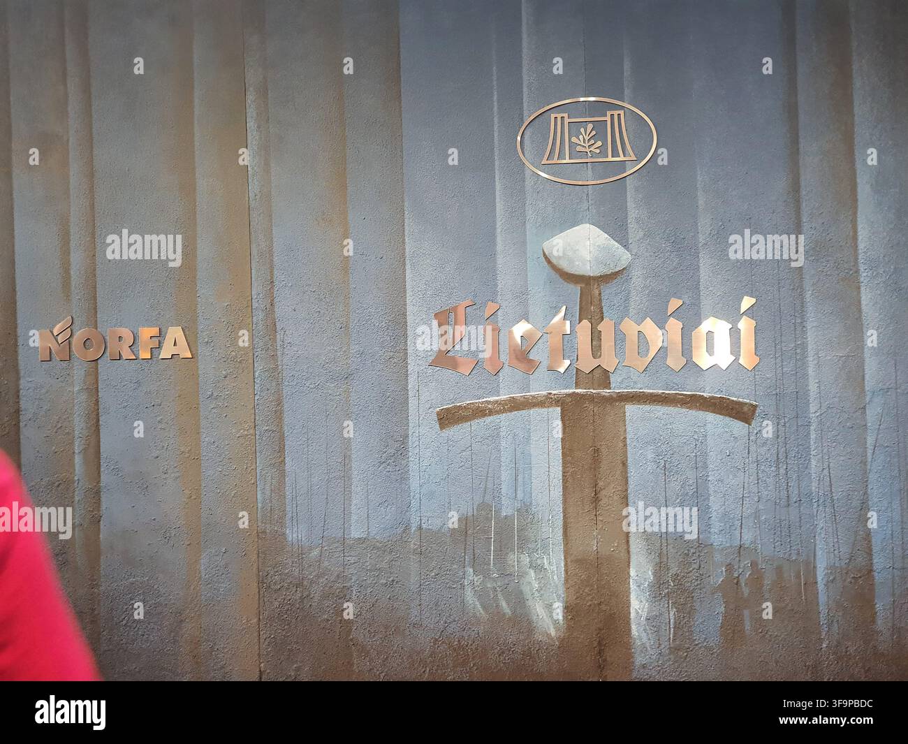 Un muro con segnaletica metallica che mostra testo e simboli lituani. Sul lato sinistro, la parola "NORFA" appare in lettere in rilievo color rame con Foto Stock
