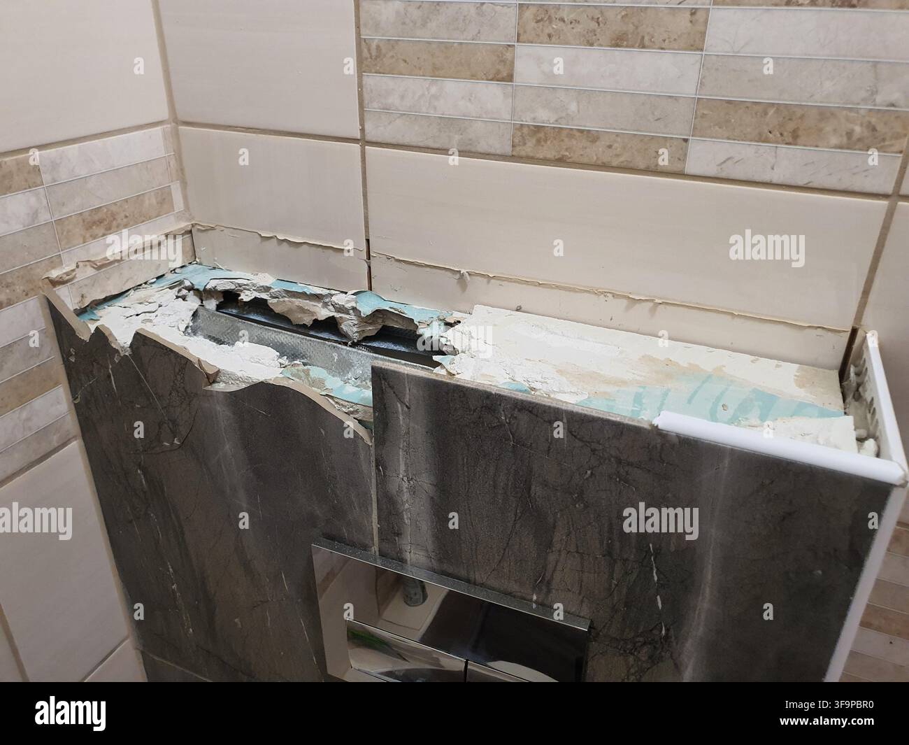 Un mobile da bagno danneggiato con un piano di lavoro distrutto in un bagno piastrellato. La superficie del piano di lavoro è rotta e si sgretola, rivelando strati Foto Stock