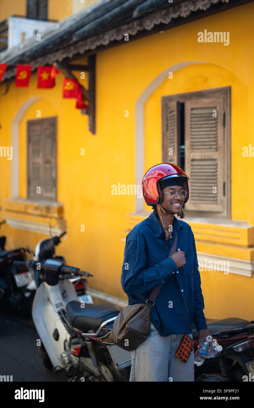 Uomo nero sorridente e allegro con casco da moto rosso che posa accanto a un caffè vintage in Vietnam. Foto Stock