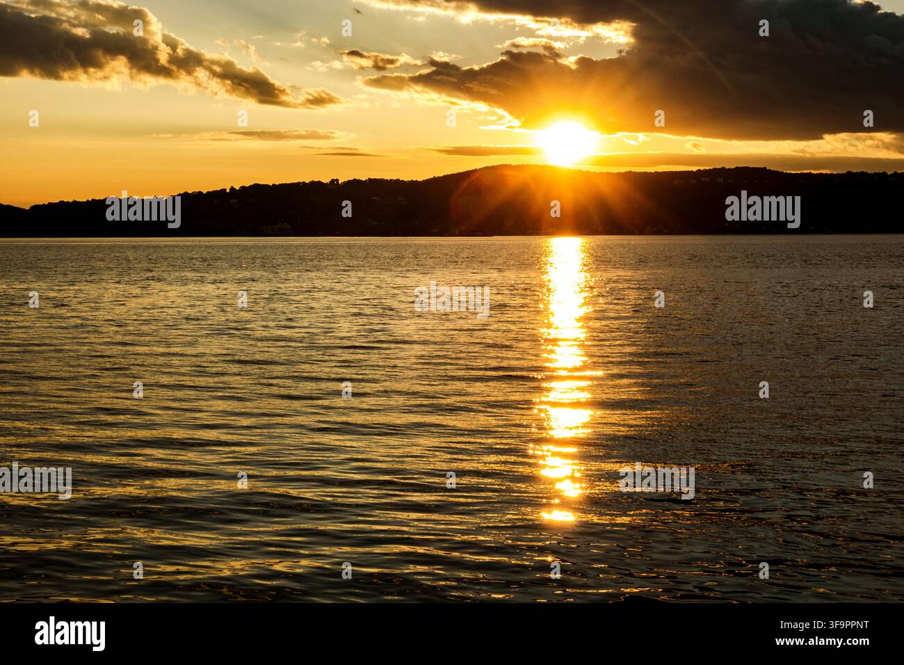 Un tramonto dorato sul fiume Hudson proietta una luce calda sull'acqua increspata, con il sole che splende sopra una riva boscosa scura e nuvole drammatiche f Foto Stock