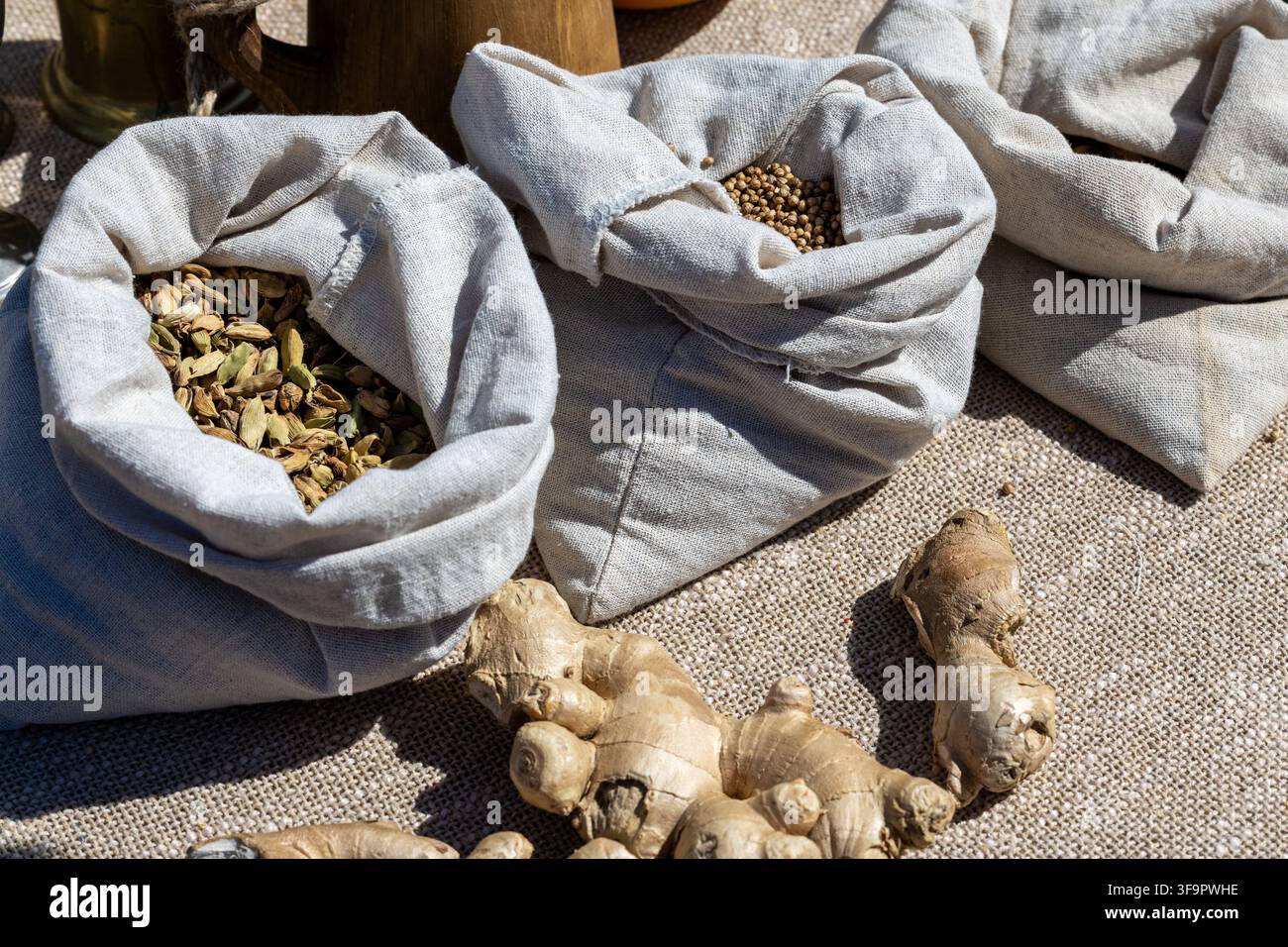 Borse di iuta contengono capsule di cardamomo essiccate e semi di coriandolo. Pezzi di zenzero fresco giacciono davanti alle borse su una superficie di tessuto intrecciato. Foto Stock