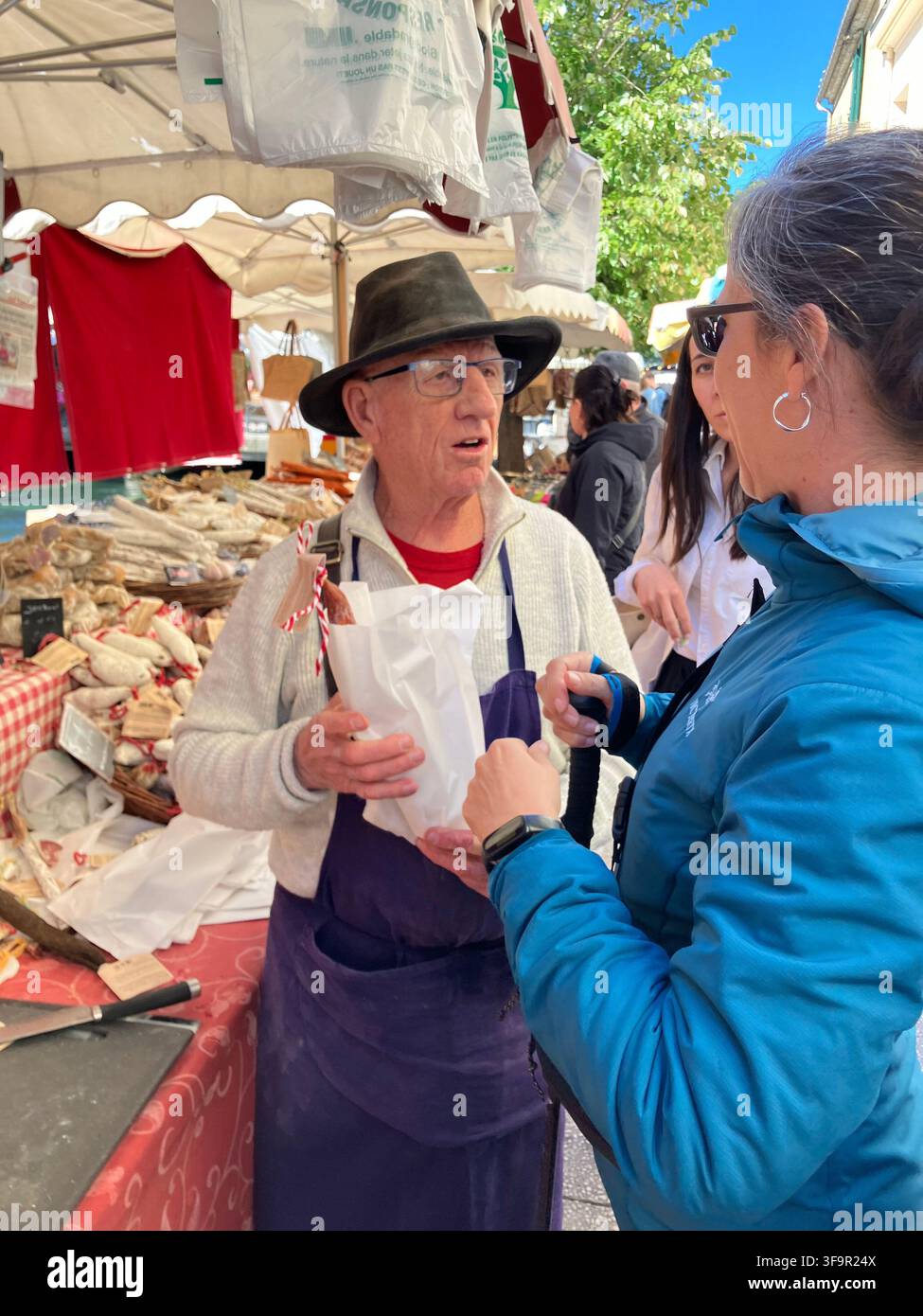 Un venditore con cappello vende salsicce essiccate locali ai clienti in un mercato di strada provenzale all'aperto a L'Isle-sur-la-Sorgue, Francia. Foto Stock