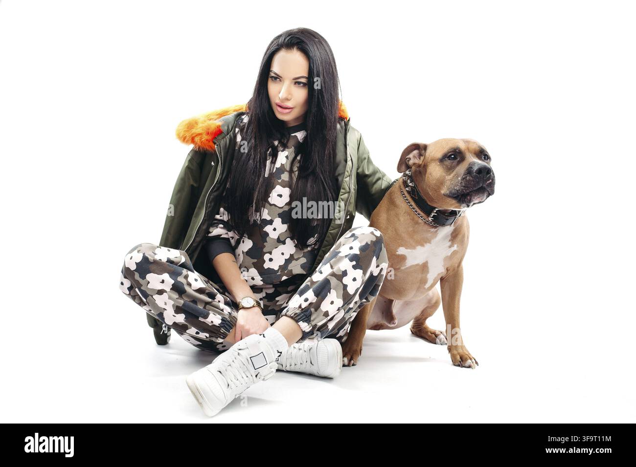 Lunghezza completa di stordimento dai capelli scuri nel modello trendy tuta sportiva e kaki giacca con arancio brillante con fodera in pelliccia sintetica tiene il cane s catena sopra backgrou bianco Foto Stock