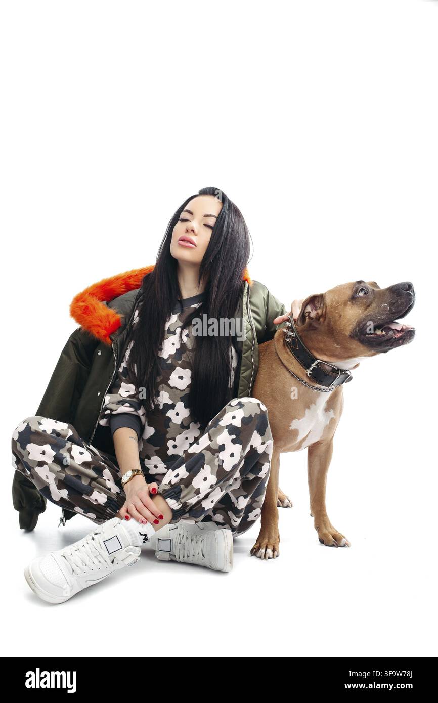 Lunghezza completa di stordimento dai capelli scuri nel modello trendy tuta sportiva e kaki giacca con arancio brillante con fodera in pelliccia sintetica tiene il cane s catena sopra backgrou bianco Foto Stock