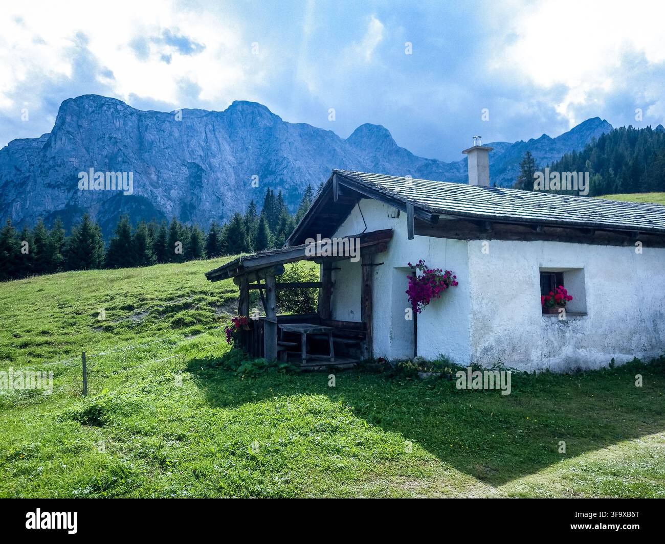 Un'incantevole capanna alpina rustica è incastonata su un prato verdeggiante, con lo sfondo di montagne maestose. Le pareti imbiancate e tradizionali w Foto Stock