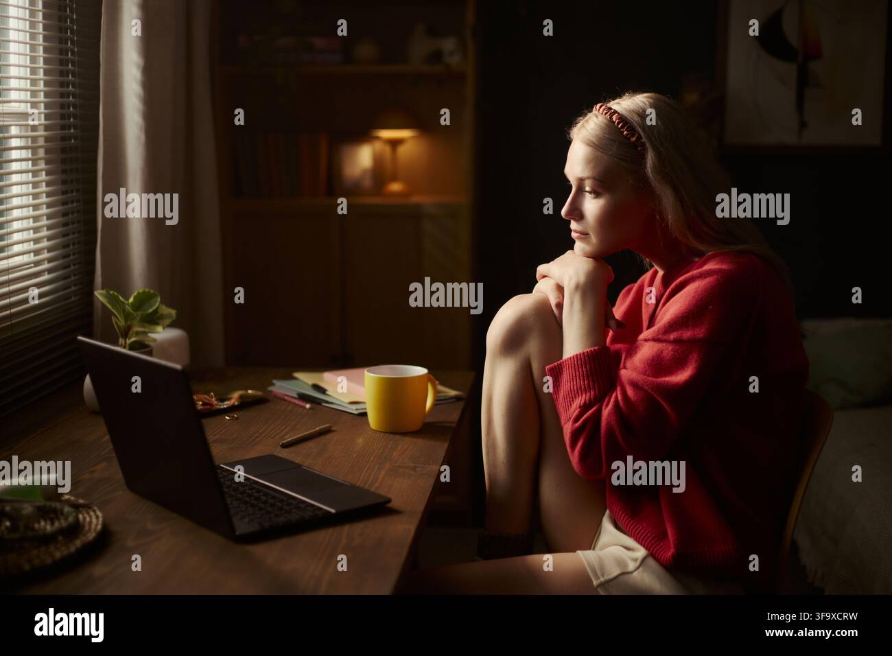 Giovane donna caucasica seduta alla scrivania, che guarda lo schermo del laptop in una stanza buia, studia o lavora da remoto. L'immagine rappresenta l'istruzione online, la stanchezza mentale e il lavoro da remoto, primo piano medio Foto Stock